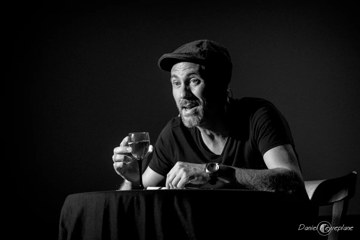 Daniel Camus attablé, casquette et verre de vin, dans le spectacle Happy Hour